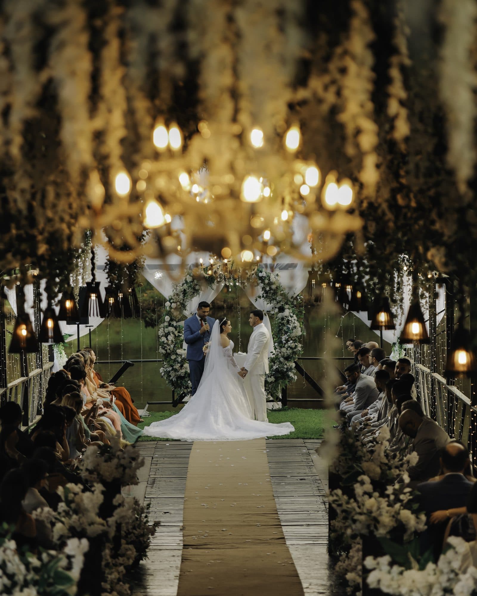 Cerimônia na ponte do Fazenda vista por entre as flores pendentes, com lustre aceso e o casal no altar