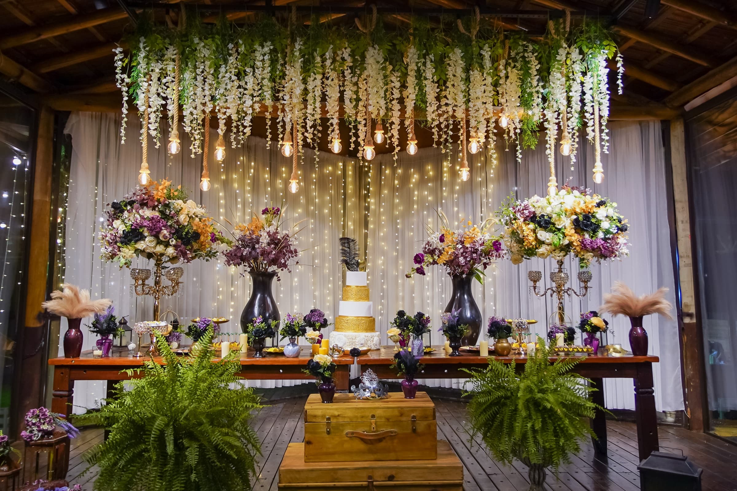 Mesa do bolo sob as pendentes de flores brancas e lâmpadas de corda