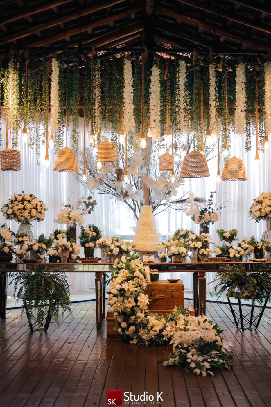 Mesa de doces com bolo e flores brancas sob luminárias de palha